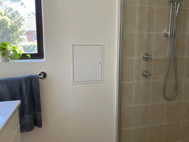 Bathroom wall with a small access panel beside a tiled shower and window in Macquarie Park